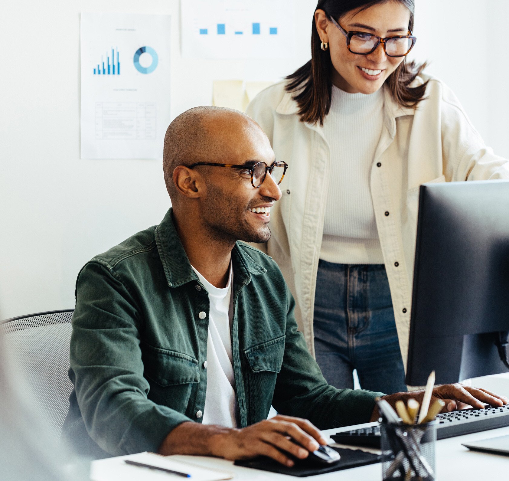 Two coworkers collaborate at a desktop workstation in a bright office. One employee sits at the computer while the other reviews information on the screen, illustrating teamwork, knowledge sharing, and data-driven decision-making in a modern workplace.